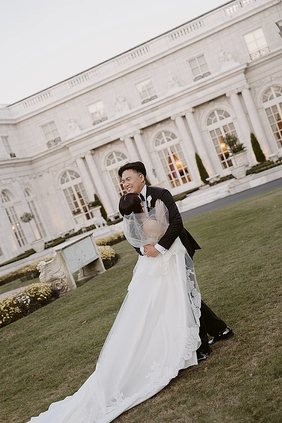 Couple portrait of bride and groom embrace, groom hugging bride as her cathedral veil blows, posed before a mansion with columns and lawn