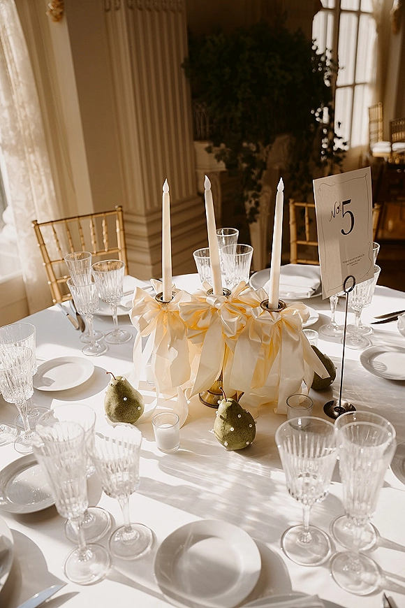 Reception tablescape wedding table setting with taper candles, ribbon bows, crystal glassware, pears, and gold chiavari chairs by sunlit windows