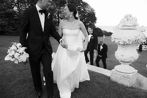 Wedding recessional as bride and groom walking hand in hand down stone steps, bride with bouquet and veil, ocean view and guests behind