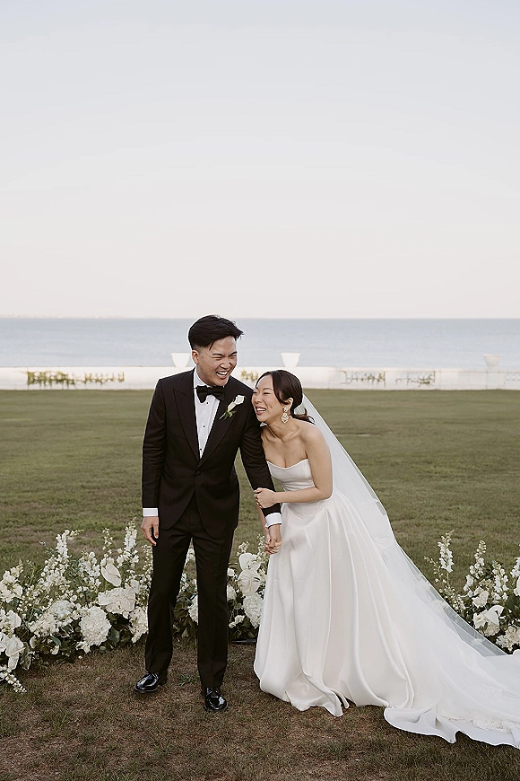Couple portrait of bride and groom laughing, holding hands on an oceanfront lawn, her veil flowing beside his black tuxedo