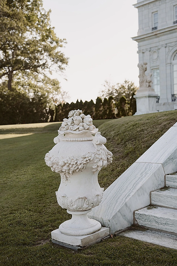 Stone garden urn at a wedding venue exterior with floral garland relief and fruit finial on marble steps by manicured lawn and hedges