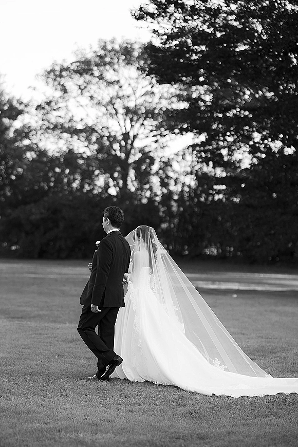 Couple portrait in black and white wedding portrait style, bride and groom walking away on a grassy field with trees and sky behind