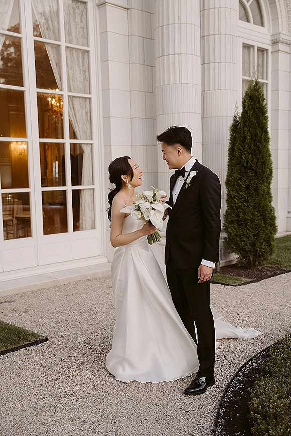 Couple portrait of bride laughing with groom, holding a calla lily bouquet outside a white building with tall windows and hedges