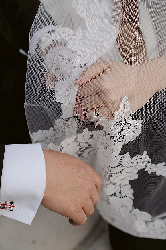 Wedding ring close-up featuring a diamond engagement ring and wedding band resting on lace veil detail against a blurred neutral background