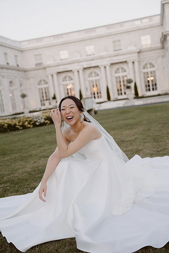 Bridal portrait of a laughing bride in a strapless wedding dress with long train and veil, seated on a lawn before a white mansion at dusk