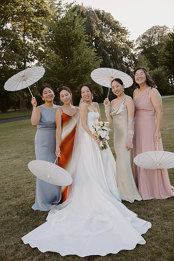 Bridesmaids photo of bride with bridesmaids holding white paper parasols, bouquet and veil on a lawn by trees at sunset