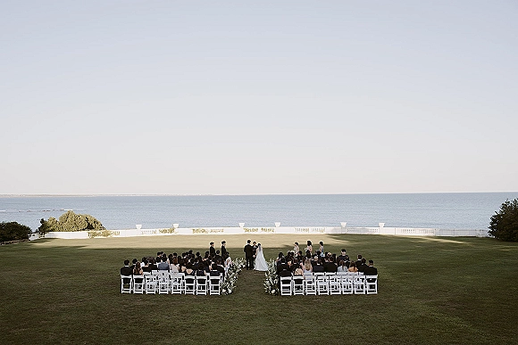 Wedding ceremony with bride and groom at the altar, veil flowing, bridesmaids and groomsmen beside them on a lawn with ocean view
