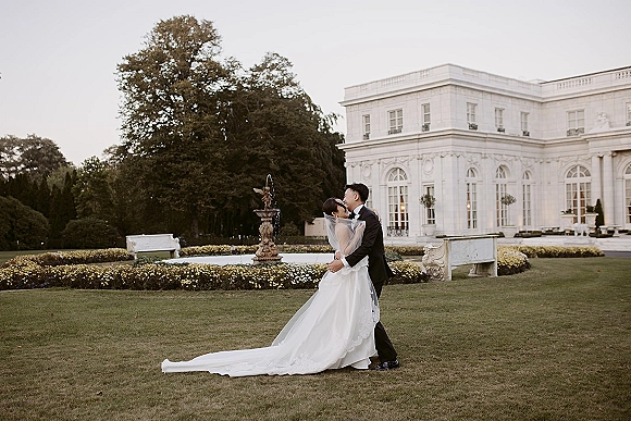 Wedding kiss portrait of bride and groom kissing in a formal garden, her long train and veil flowing near a fountain and estate building