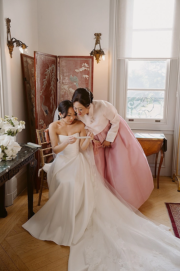 Bride with mother in a pink hanbok helping bride adjust veil and lace train in a bright bridal suite with window light and folding screen