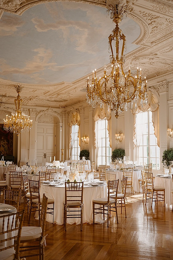 Reception tablescape with round wedding reception tables, white linens, taper candles and gold chiavari chairs beneath chandeliers in an ornate ballroom
