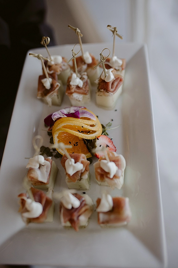 Cocktail hour appetizers on a white platter with prosciutto and cheese cubes on skewers, topped with microgreens, on a white tablecloth indoors