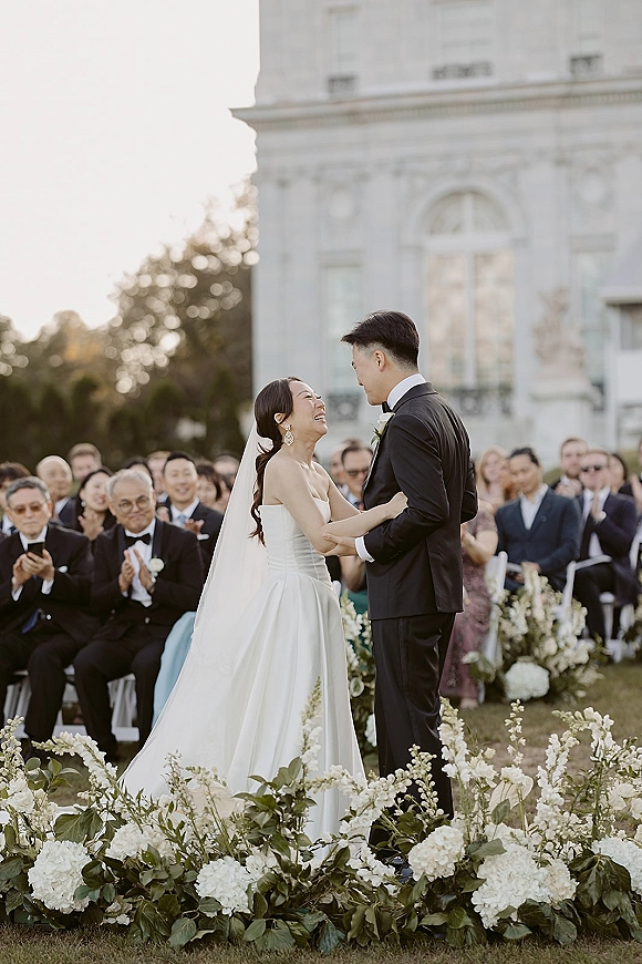 Ceremony moment at an outdoor wedding ceremony as bride and groom hold hands and laugh, framed by white hydrangea aisle florals and guests on a lawn