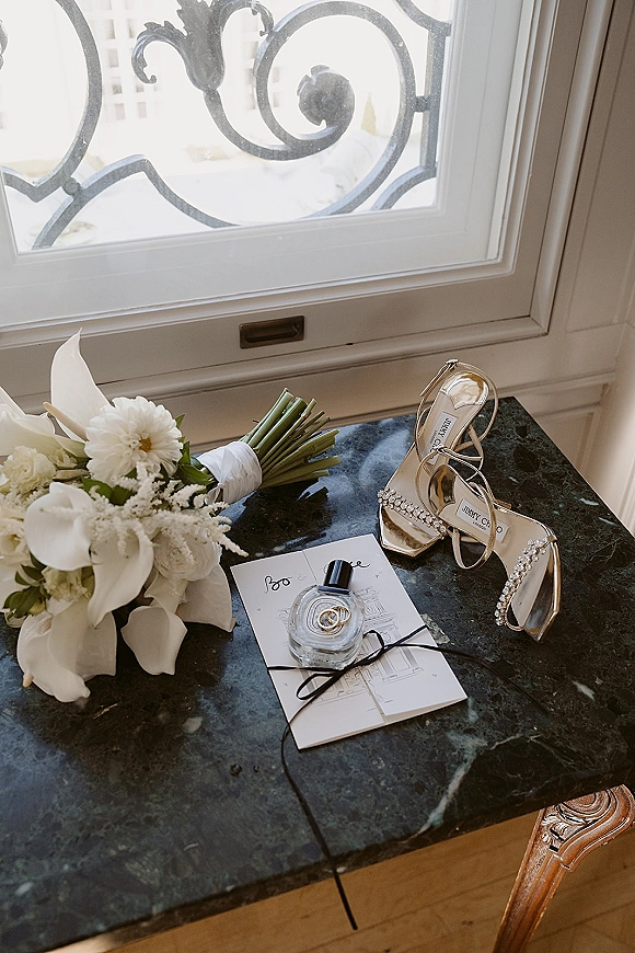 Wedding details flatlay with bridal accessories on marble, featuring calla lily bouquet, gold strappy heels, rings on invitation, perfume by window