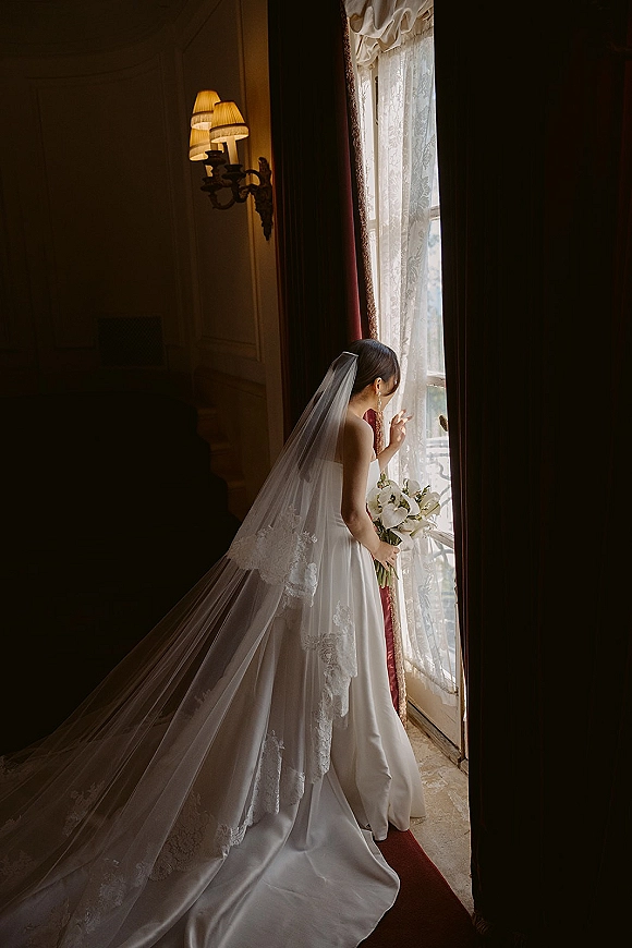 Bridal portrait of a bride looking out window, holding calla lily bouquet with long lace train in soft window light by drapes