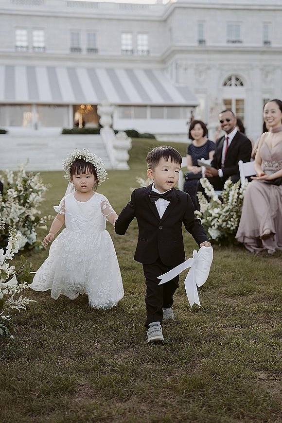 Flower girl and ring bearer walk the aisle, ring bearer holding pillow, as guests watch on an outdoor lawn with white chairs and greenery
