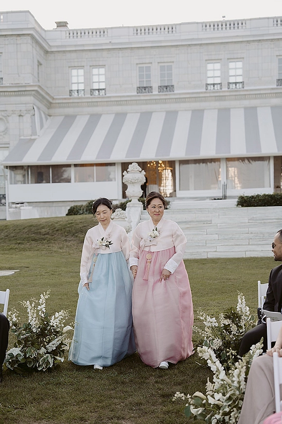 Wedding processional with parents walking down aisle in hanbok and corsages, passing white aisle flowers and greenery on a lawn by stone steps