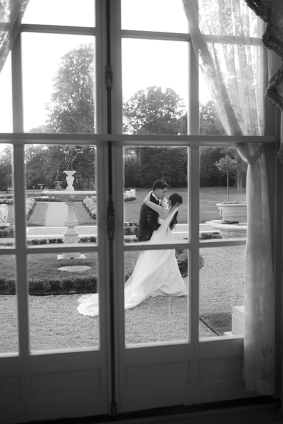 Wedding kiss portrait in black and white, bride and groom kissing framed by french doors, her veil and dress train flowing toward the garden fountain