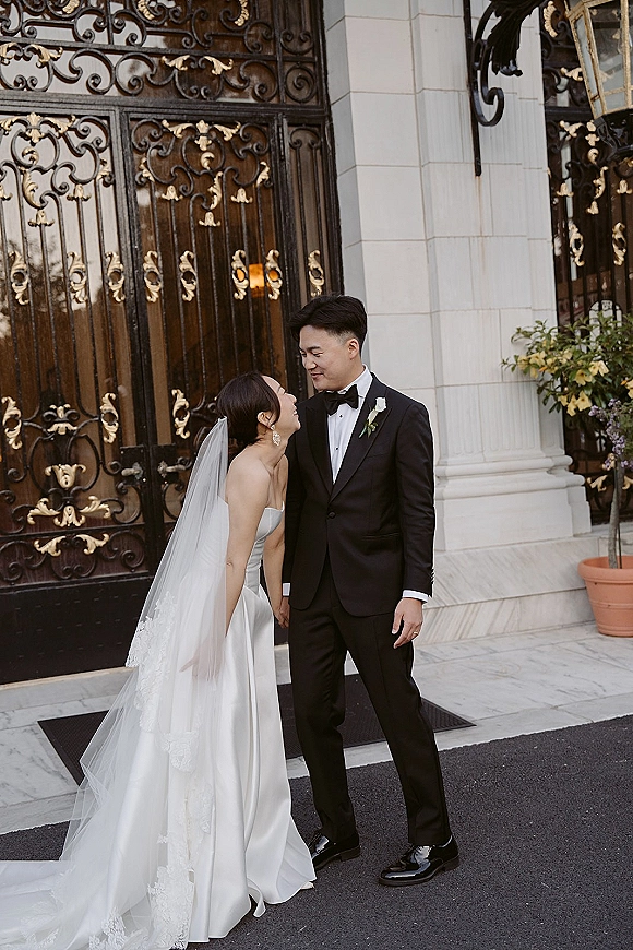 Couple portrait of bride looking up at groom, her veil draping over a satin dress, beside ornate wrought iron doors and stone columns