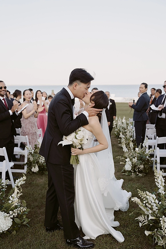 Wedding kiss at an oceanfront ceremony as bride in strapless satin dress and veil holds white bouquet while guests watch along floral aisle