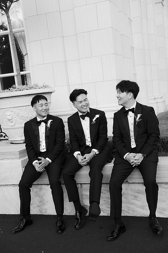Groomsmen portrait of three men in black tuxedos and bow ties laughing on stone steps outside a building, boutonniere accents visible