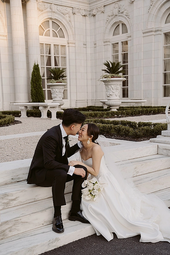 Couple portrait of bride and groom sitting on steps, sharing a forehead kiss; strapless dress and veil against marble building facade