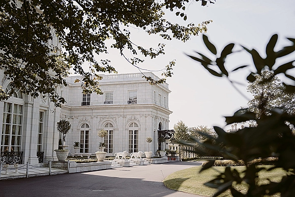 Wedding venue exterior with white stone facade, arched windows and French doors, framed by potted topiary trees along a paved driveway