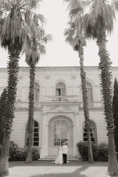Couple portrait rendered as a black and white wedding portrait before a historic mansion facade with palm trees, bride in a classic bridal gown.