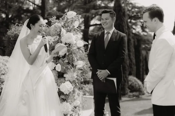Ceremony moment with the bride reading vows from a vow book, beside the groom and officiant, set amid garden trees, hedges, and a lawn.