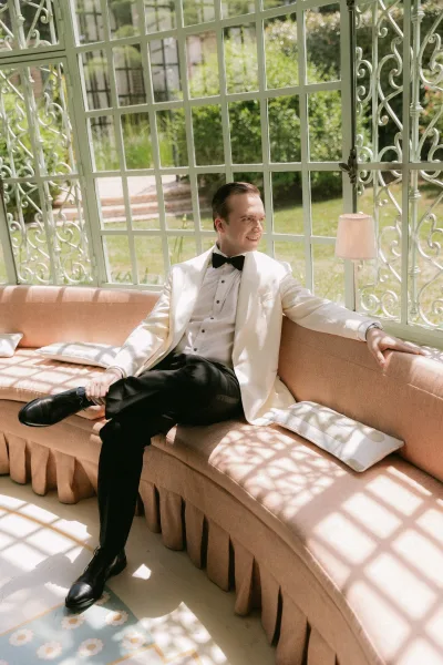 Groom portrait of a groom in a white dinner jacket, neatly poised by wrought iron windows, his black bow tie crisp against the jacket.