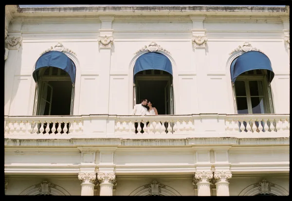 Wedding kiss portrait of a couple at the balcony railing, a balcony wedding photo set against a historic facade with arched windows and blue awnings.