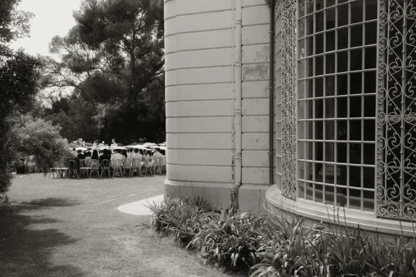 Ceremony moment during an outdoor garden ceremony, guests shaded by parasols beneath trees by a historic facade with wrought iron windows