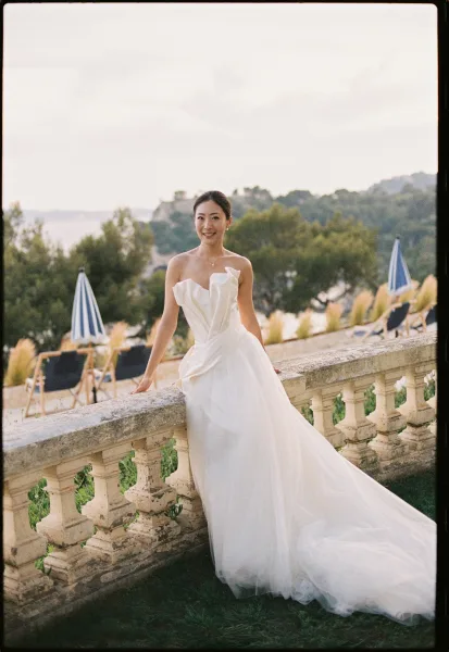 Bridal portrait in a strapless wedding dress with a long train, posed by a stone balustrade overlooking a garden and water with distant hills.
