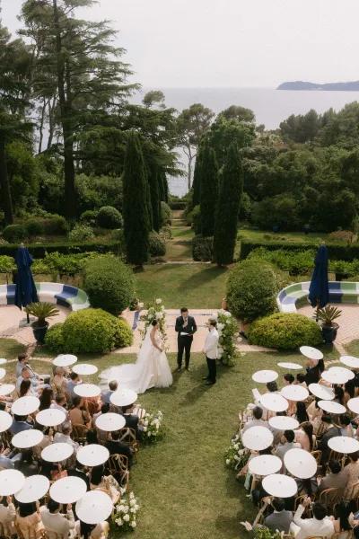 Ceremony moment during an outdoor garden ceremony as the bride and groom stand at the altar, white parasols dotting the seating, with ocean beyond.
