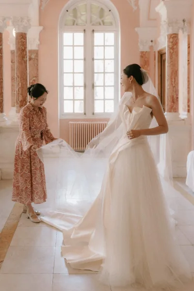 Bride getting ready as someone is helping with her bridal veil, bow-detail bodice straightened by a large window in a pink-walled room.