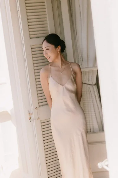Bridal portrait of a bride by a window, her satin slip dress catching soft light as she turns in profile against shutters and a white wall.
