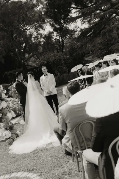 Ceremony moment during wedding vows beneath garden trees, the bride's long tulle veil catching light as guests watch on the lawn.