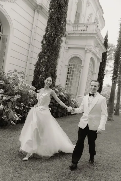 Bride and groom walking, couple holding hands across a grass lawn, the groom in a black bow tie, with a stately mansion and garden shrubs behind.