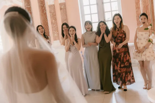 Bridesmaid first look during a bride reveal moment as the bride in a bridal veil faces cheering bridesmaids amid marble columns and tall windows.