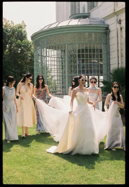 Bride with bridesmaids, a bridal party walking across a lawn as they lift her tulle train by a wrought-iron gazebo and historic estate facade