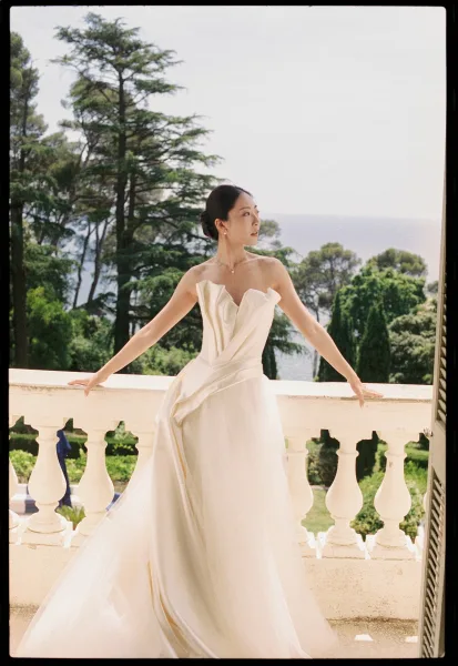 Bridal portrait of a bride on balcony in an ivory strapless gown, sleek low bun, framed by a stone balustrade with ocean and sky beyond.