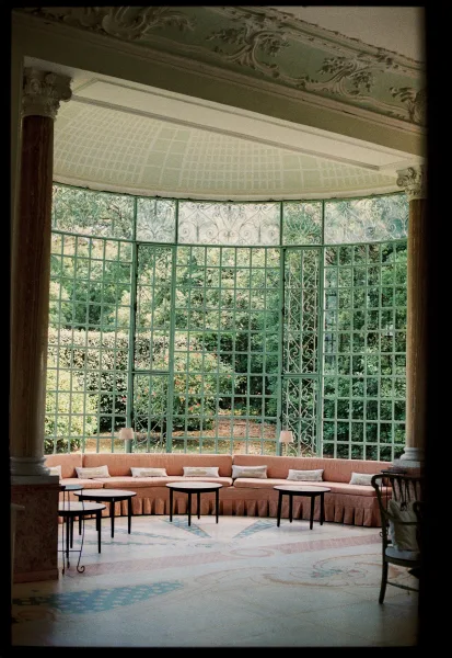 Wedding lounge arranged for cocktail hour seating, featuring a curved banquette beneath tall ironwork windows overlooking garden trees.