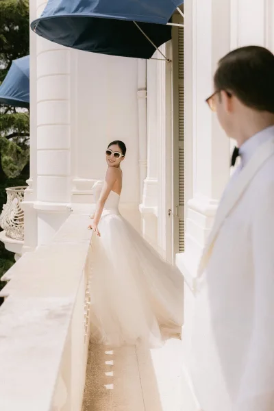 Bridal portrait of a bride on balcony, wearing chic sunglasses, framed by white columns and blue awnings with greenery beyond.
