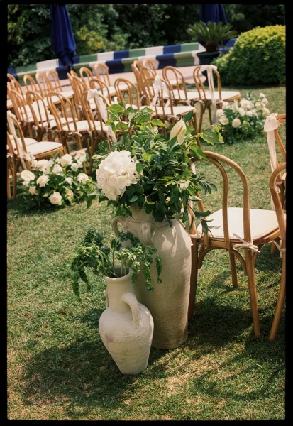 Ceremony aisle decor with white roses anchors a garden ceremony setup on a grass lawn, with garden shrubs and a striped bench in the background.