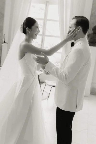First look moment and groom's reaction as the bride touches his shoulder, her veil trailing by a window with sheer curtains