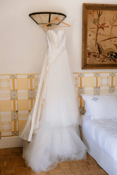 Wedding dress, a tulle skirt wedding dress, hangs in a bedroom with a patterned wall, framed artwork, and a bed with pillows.