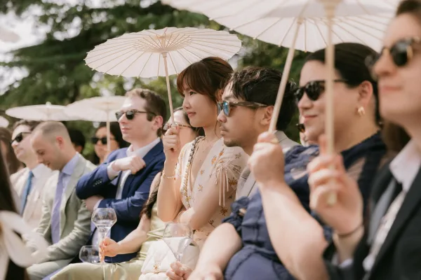 Ceremony guests, wedding guests with parasols, sit as paper parasols cast shade amid garden greenery during a sunny outdoor ceremony.