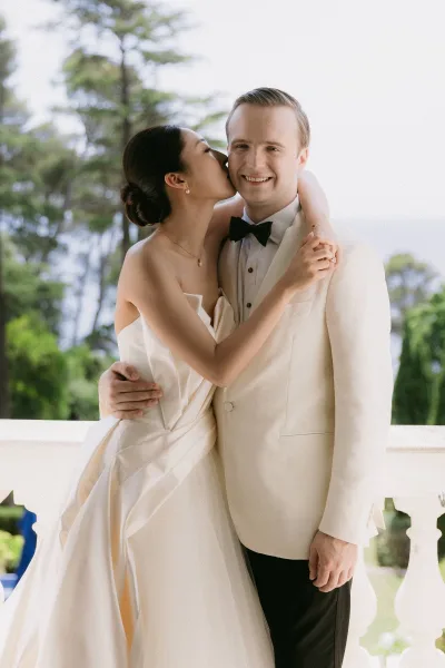 Couple portrait, bride kissing groom on a balcony, wedding bands gleaming; garden trees and distant water shimmer beneath the sky.