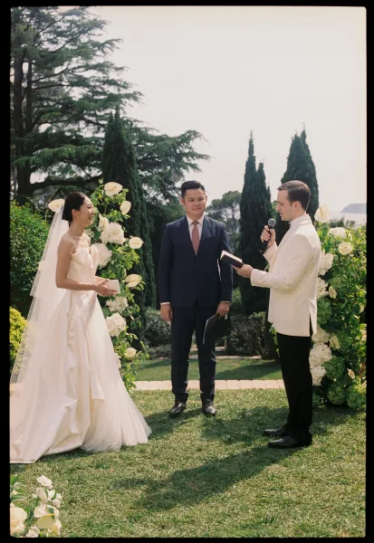 Wedding vows during an outdoor vow exchange, the groom reading vows to his smiling bride while holding a microphone before the officiant.