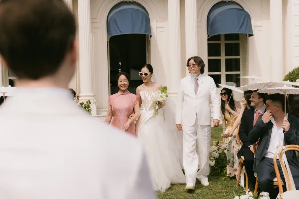 Ceremony processional with the bride walking down the aisle, guests shaded by paper parasols on a lawn before a columned building with blue awnings.