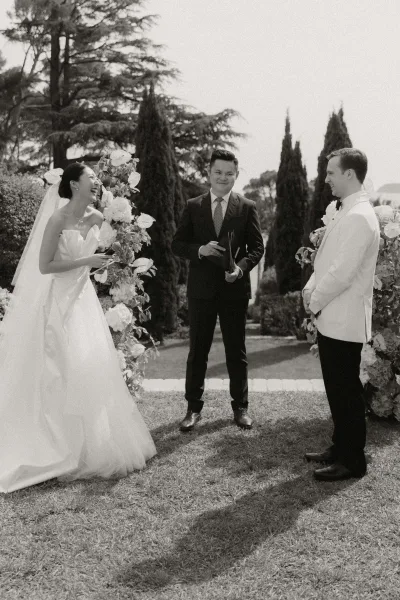 Ceremony moment as the bride laughs during outdoor wedding vows, holding a microphone while facing the groom by lush garden hedges.